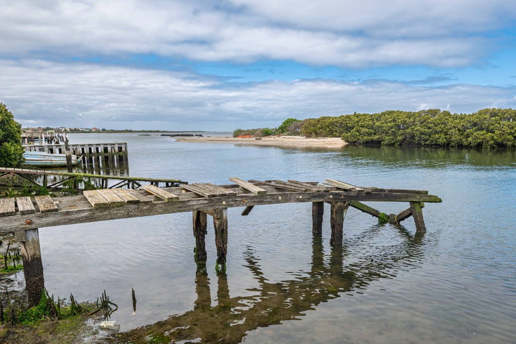 Weathered wooden pier extending over calm waters with a mangrove backdrop and cloudy sky.