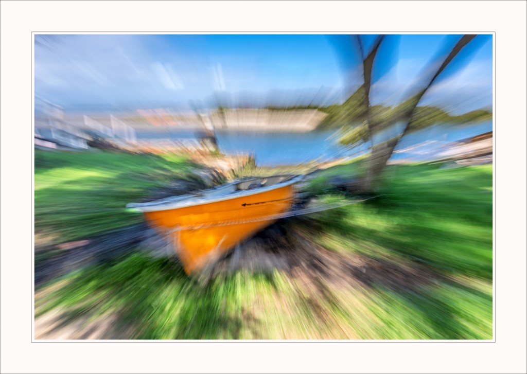 A blurred image of an orange boat on grassy land with a background of water and trees.